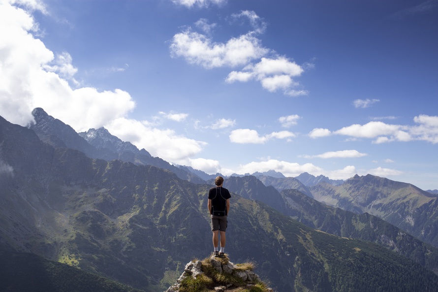 Man in Black Shirt and Gray Shorts Standing on Cliff Under White and Blue Cloudy Sky