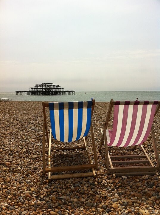 Hier sind zwei leere Liegestühle zu sehen, die auf dem Kieselstrand Brightons stehen und gen Meer gerichtet sind